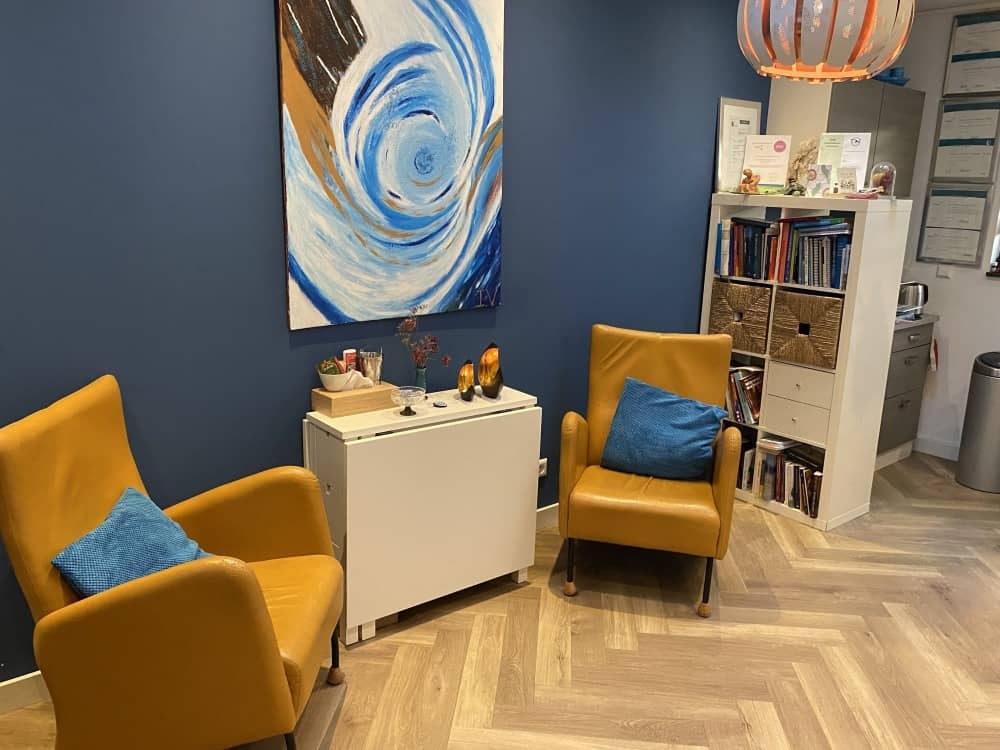 Praktijkruimte Chakora omgeving Brabant - Ingericht met twee gele stoelen, een blauwe muur met kunstwerk en een boekenkast